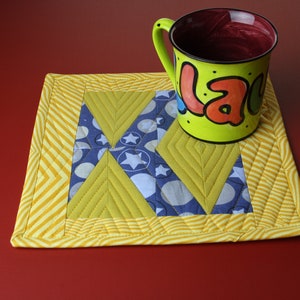 Könnte beinhalten: Ein gelber, gesteppter Untersetzer mit geometrischem Design, mit blauen und weißen Stern- und Kreismustern. Ein limettengrüner Becher mit dem Wort "Lala" in bunten Buchstaben steht auf dem Untersetzer. Der Becher hat ein rotes Innenleben.