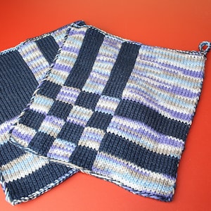 Könnte beinhalten: Zwei gehäkelte Topflappen in einem gestreiften Muster in Blau, Weiß und Lila. Die Topflappen sind aus dickem Garn gefertigt und haben eine Schlaufe zum Aufhängen.