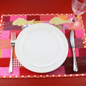 May include: A white dinner plate and silver cutlery are set on a red and white patchwork placemat. The placemat has a white polka dot border.