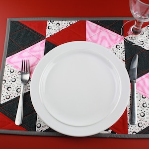 Op de afbeelding: Een wit dinerbord met een zilveren vork en mes op een grijs en wit gestikt placemat met roze, rode en zwarte geometrische patronen.