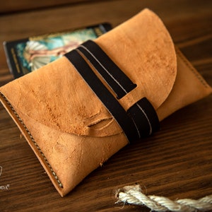 May include: A light brown leather pouch with a black leather strap. The pouch is closed with a flap and has a stitched edge. The pouch is sitting on a wooden surface.