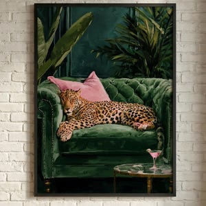 May include: A framed wall art print featuring a leopard resting on a deep green tufted velvet sofa. A soft pink pillow sits behind the leopard, and a small round table with a pink cocktail glass is positioned in front of the sofa.