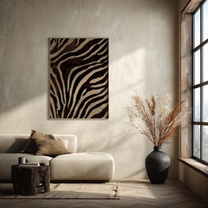 May include: A rectangular wall art piece featuring a black and beige zebra stripe pattern in a thin frame. The artwork is displayed on a light beige wall above a cream-coloured sofa with brown cushions, next to a black vase with dried pampas grass.
