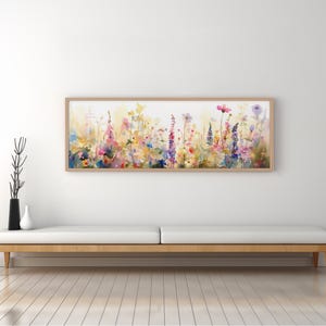 Puede incluir: Una pintura de acuarela de un campo de flores silvestres en tonos de rosa, morado, amarillo y blanco. Las flores están dispuestas en una línea horizontal a través del lienzo, creando una sensación de movimiento y energía. La pintura está enmarcada en un marco de madera claro.