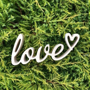 Peut inclure: Découpe en bois blanc du mot "love" avec une forme de cœur à la fin du "e". La découpe est placée sur un lit de feuillage vert.