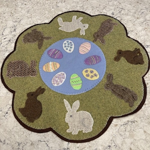 May include: A green felt Easter bunny doormat with a blue circle in the center. The circle is decorated with colorful embroidered eggs and surrounded by brown and white felt bunnies.