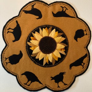 May include: A brown felt wall hanging with a black stitched border. The centre features a yellow and brown sunflower with a black centre. Eight black crow silhouettes are stitched around the sunflower.