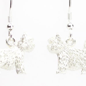 May include: A pair of silver-colored dog-shaped earrings. Each earring features a detailed terrier design, with a textured surface and a hook for pierced ears. The earrings are displayed against a white background.