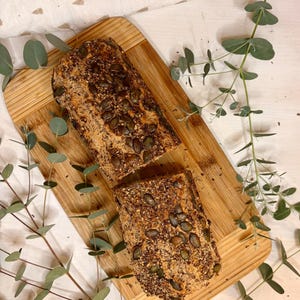 May include: A sliced loaf of bread on a wooden board, scattered with seeds. The bread is surrounded by sprigs of eucalyptus. The background includes a white cloth with a subtle pattern.