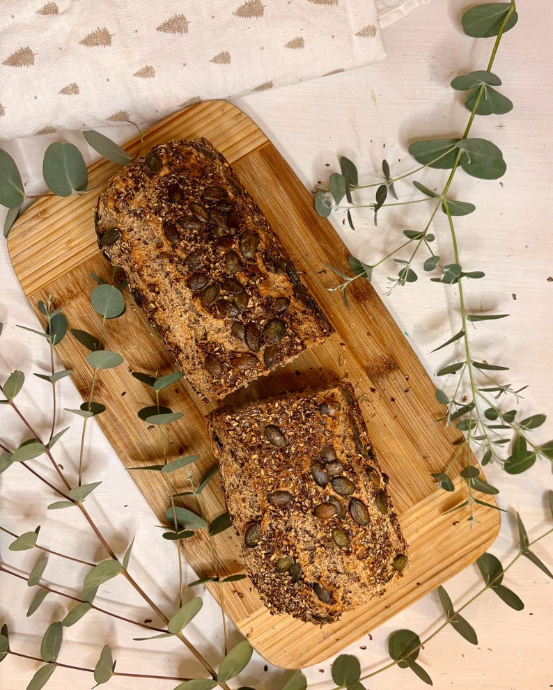 May include: A sliced loaf of bread on a wooden board, scattered with seeds. The bread is surrounded by sprigs of eucalyptus. The background includes a white cloth with a subtle pattern.