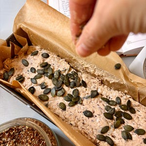 May include: A loaf of bread in a baking tin, lined with parchment paper, being sprinkled with seeds. The bread is speckled with seeds and grains, and topped with large green pumpkin seeds. A jar of seeds and a wooden spoon are also visible.