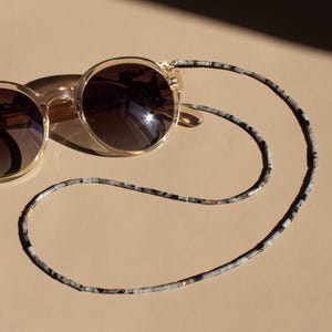 Op de afbeelding: Ronde zonnebril met een transparant montuur en donkere glazen, gecombineerd met een brillenkoord met patroon. De ketting heeft een zwart-wit gespikkeld ontwerp. De zonnebril ligt op een beige oppervlak, met schaduwen.