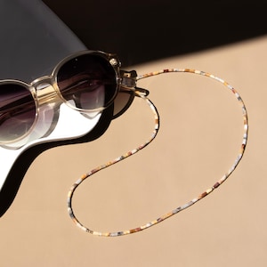 May include: Round sunglasses with translucent frames and dark gradient lenses attached to a long, thin beaded chain. The chain features small, rectangular beads in a mottled pattern of brown, white, tan, and grey tones, creating a continuous loop.