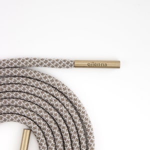 May include: A coiled shoelace with a brown and white checkered pattern. The shoelace has gold-colored aglets and the word "eilenna" is printed on one of the aglets. The shoelace is on a white background.