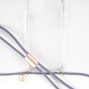 Puede incluir: Una funda de tel&eacute;fono transparente con una correa de tel&eacute;fono gris unida. La correa tiene un acento dorado y un peque&ntilde;o dije dorado con una letra.