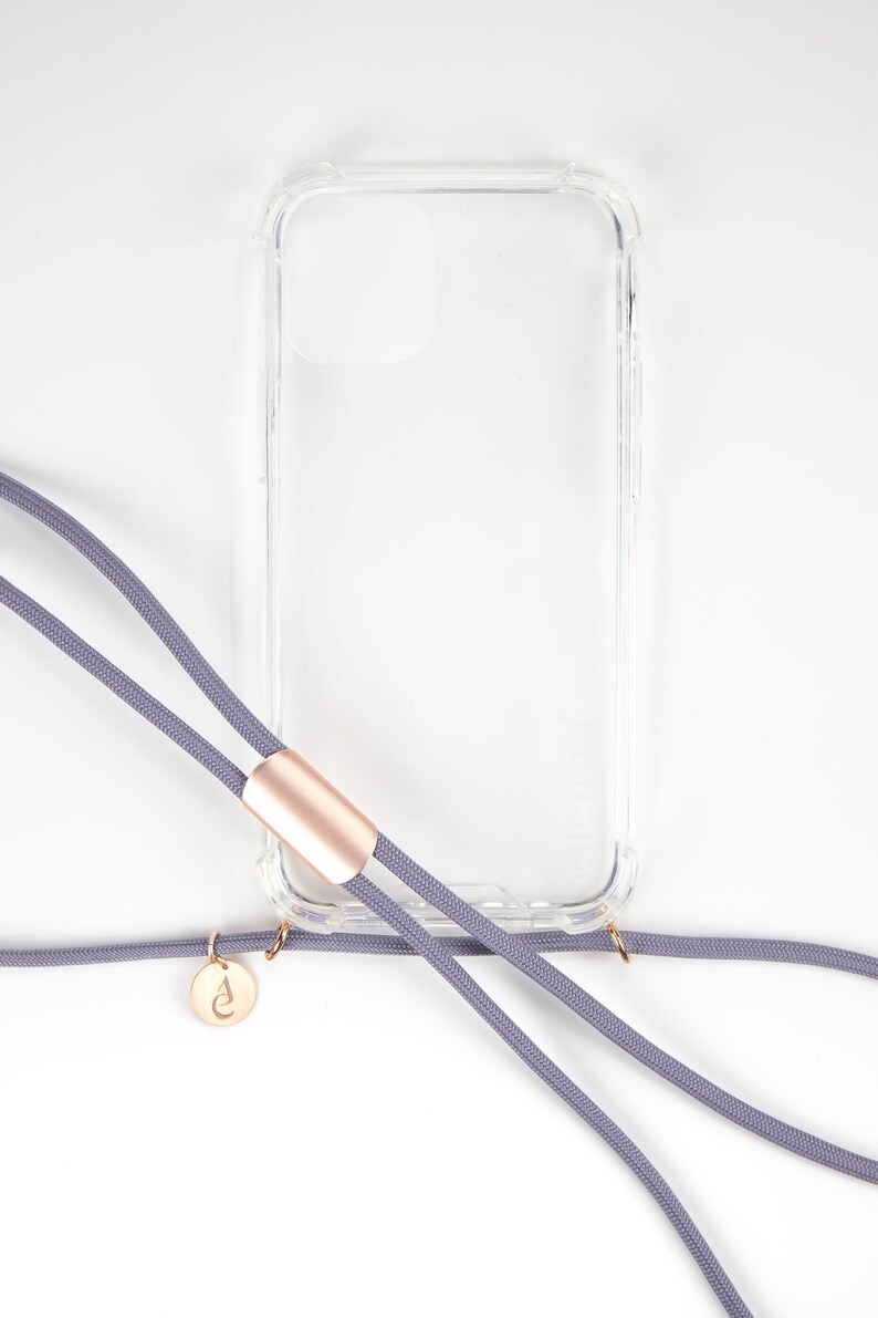 Puede incluir: Una funda de tel&eacute;fono transparente con una correa de tel&eacute;fono gris unida. La correa tiene un acento dorado y un peque&ntilde;o dije dorado con una letra.