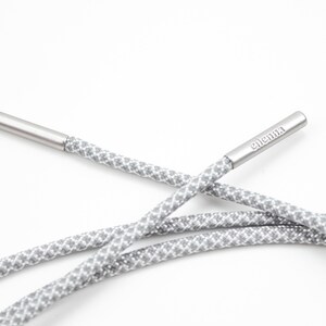 May include: Gray and white patterned shoelaces with silver aglets. The aglets are engraved with the word "elementa".