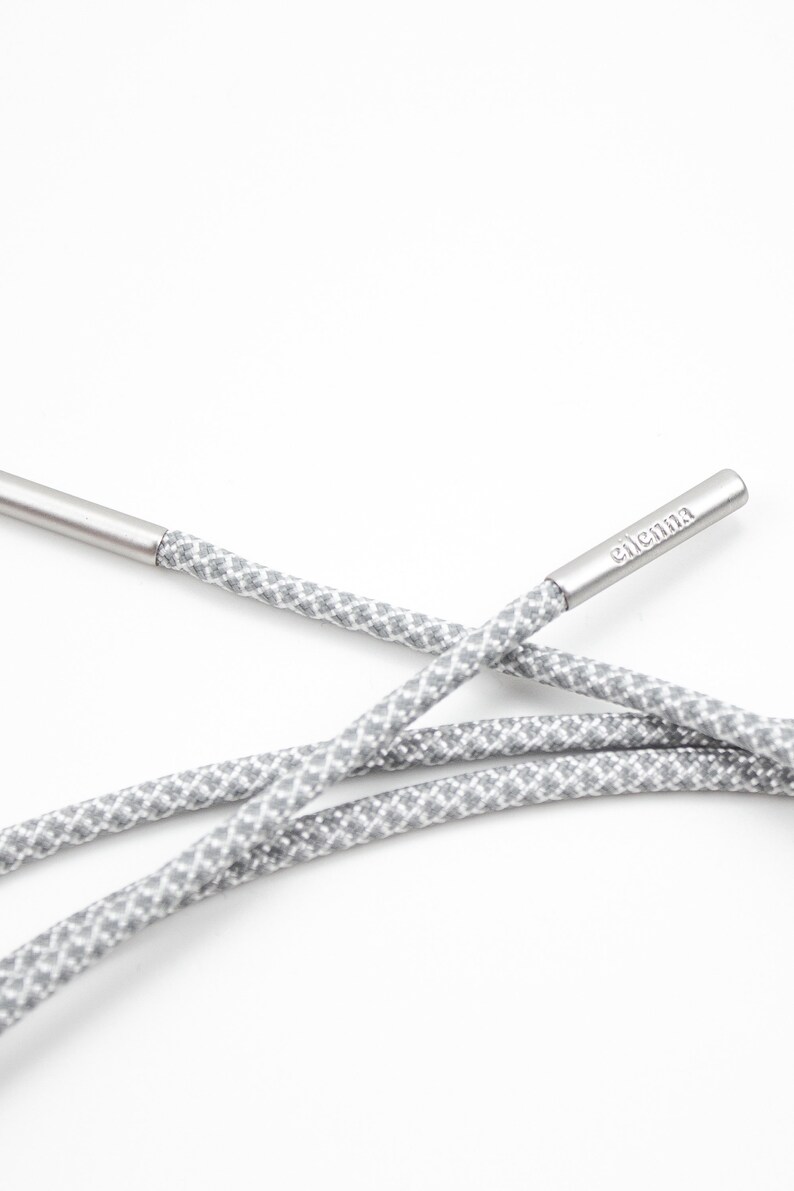 May include: Gray and white patterned shoelaces with silver aglets. The aglets are engraved with the word "elementa".