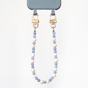 Peut inclure: Une coque de smartphone bleue avec le mot eilenna embossé au dos, fixée à une dragonne perlée. La dragonne présente des perles sphériques alternées bleu clair et crème, reliées par des fermoirs mousquetons en métal doré.