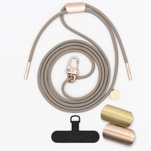 以下が含まれることがあります： ローズゴールドのアクセントが付いたグレーの電話用ストラップ。カラビナ、エンドキャップ、小さなチャームが含まれています。黒い電話タブと、金とローズゴールドの2つの金属シリンダーが含まれています。