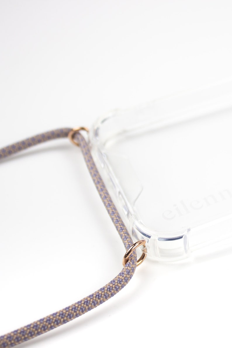 May include: A clear phone case with a gray and beige braided strap attached to the case with gold rings. The case has the word "eleni" printed on the back.