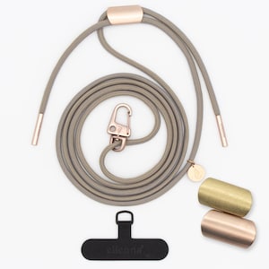 Può includere: Un cordino grigio per telefono con dettagli oro rosa, tra cui una chiusura, estremità del cordino e un piccolo ciondolo. Il set include due pezzi metallici cilindrici, uno dorato e uno oro rosa, e un adattatore per telefono nero con il nome del marchio.