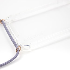 Puede incluir: Una funda transparente para tel&eacute;fono con un cord&oacute;n gris adjunto. La funda tiene un borde elevado alrededor de la pantalla y la lente de la c&aacute;mara. El cord&oacute;n tiene dos anillos dorados que se sujetan a la funda.