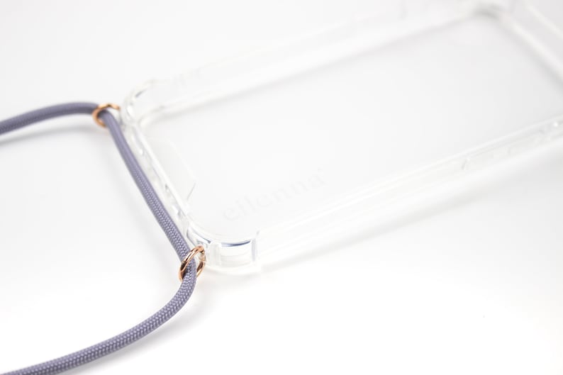 Puede incluir: Una funda transparente para tel&eacute;fono con un cord&oacute;n gris adjunto. La funda tiene un borde elevado alrededor de la pantalla y la lente de la c&aacute;mara. El cord&oacute;n tiene dos anillos dorados que se sujetan a la funda.