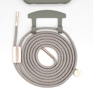 May include: A coiled gray and white patterned phone strap with rose gold hardware and a matching phone case. The strap has a circular design with a rose gold clasp and a small, round charm. The phone case and strap are olive green.