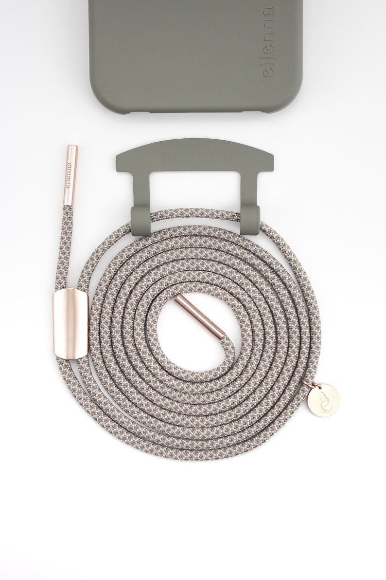 May include: A coiled gray and white patterned phone strap with rose gold hardware and a matching phone case. The strap has a circular design with a rose gold clasp and a small, round charm. The phone case and strap are olive green.