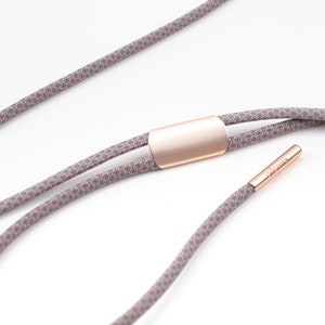 May include: A double-strand cord necklace with a rose gold-toned metal clasp and a small rose gold-toned tag with the word "eleonna" engraved on it. The cord is a light brown color with a subtle purple pattern.