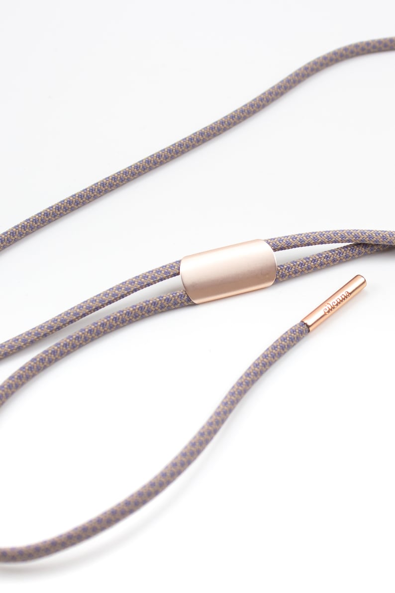 May include: A double-strand cord necklace with a rose gold-toned metal clasp and a small rose gold-toned tag with the word "eleonna" engraved on it. The cord is a light brown color with a subtle purple pattern.