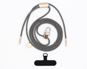 May include: A black and white patterned phone lanyard with rose gold hardware. The lanyard includes a clasp, a small charm, and a black phone connector with the brand name. The lanyard is designed to be worn around the neck.