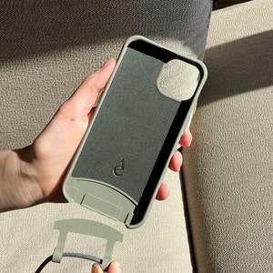 May include: A sage green phone case with a matching strap attachment. The case has a dark gray interior and a small logo. The strap attachment is connected to a black cord. The brand name "elienna" is printed on the attachment.