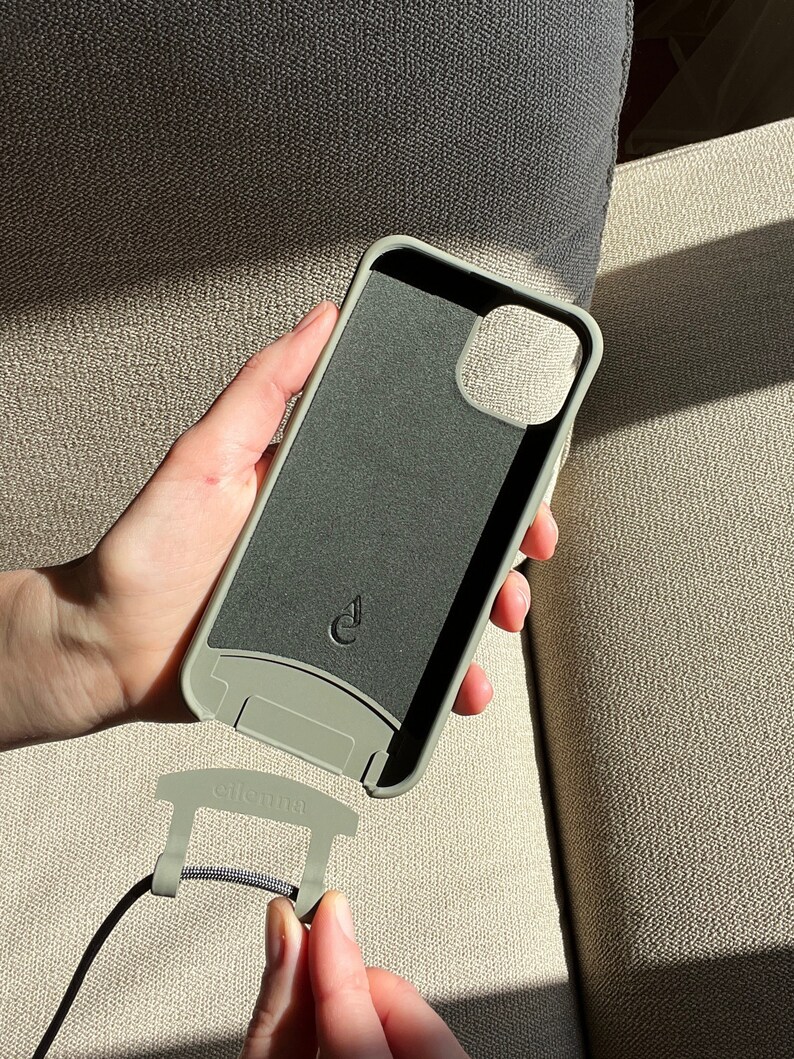 May include: A sage green phone case with a matching strap attachment. The case has a dark gray interior and a small logo. The strap attachment is connected to a black cord. The brand name "elienna" is printed on the attachment.