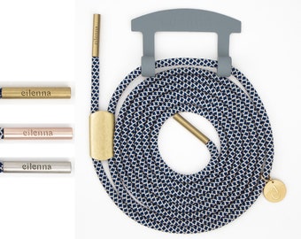May include: A navy blue and white patterned shoelace with a gold metal tip and a gold metal charm with the letter "C" on it. The shoelace is coiled and hanging from a gray metal hook.