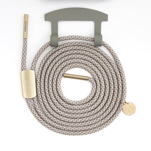 May include: A phone case and lanyard set in neutral tones. The olive green case and holder are paired with a beige and brown patterned rope lanyard. The lanyard features gold-colored metal accents and a small charm.