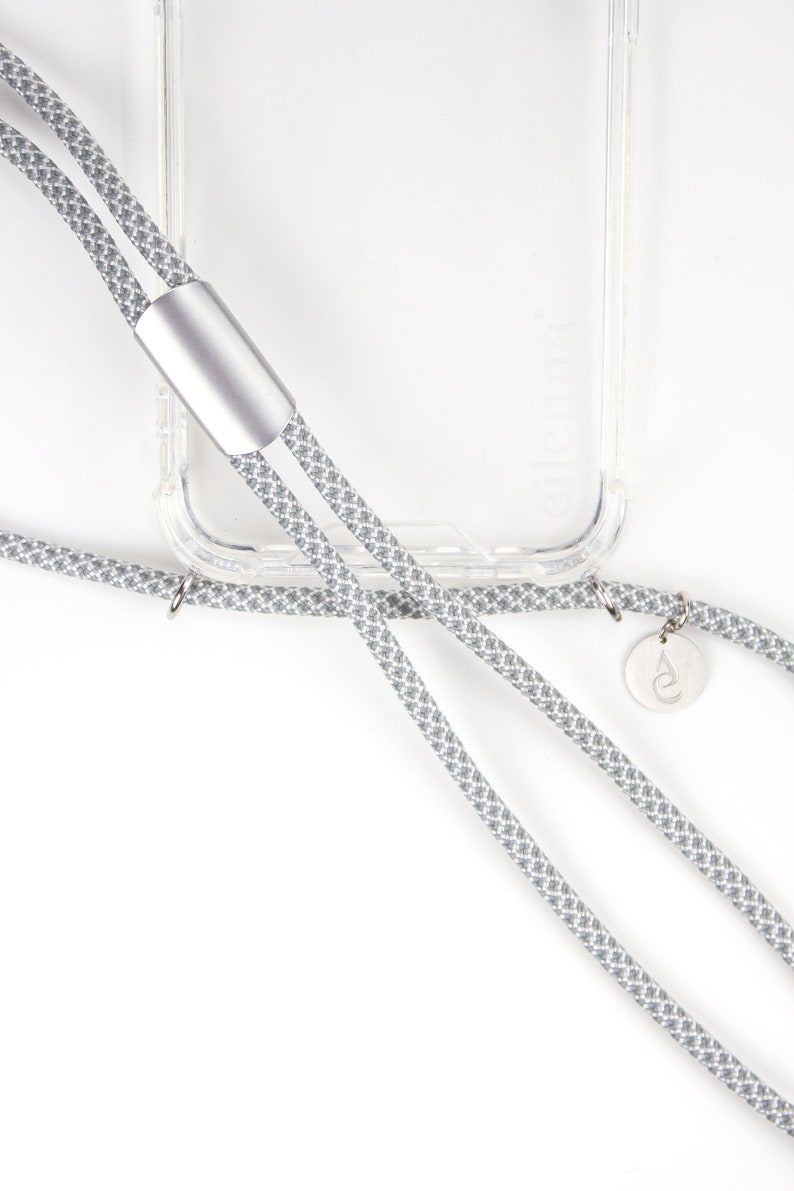 May include: A gray and white patterned phone strap with a silver accent and a small silver charm. The strap is attached to a clear phone case.