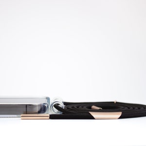May include: A clear phone case with a black phone lanyard. The lanyard has rose gold hardware.