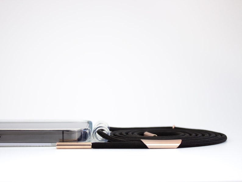 May include: A clear phone case with a black phone lanyard. The lanyard has rose gold hardware.