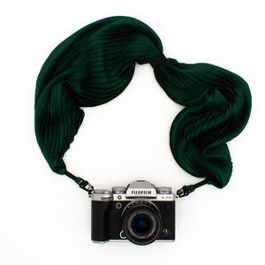 Może przedstawiać: Czarny i srebrny aparat Fujifilm X-T5 z ciemnozielonym, plisowanym paskiem do aparatu. Pasek do aparatu jest zawinięty wokół aparatu i ma czarne złącza. Aparat jest wyśrodkowany na białym tle.