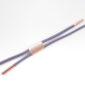 Puede incluir: Un par de cordones grises con puntas de metal dorado y un deslizador de metal dorado. Los cordones est&aacute;n dispuestos en plano sobre un fondo blanco.