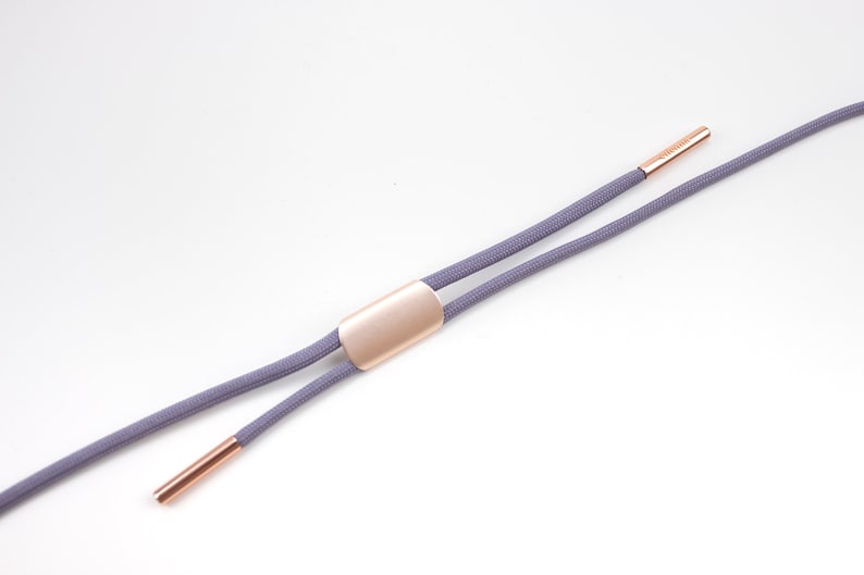 Puede incluir: Un par de cordones grises con puntas de metal dorado y un deslizador de metal dorado. Los cordones est&aacute;n dispuestos en plano sobre un fondo blanco.