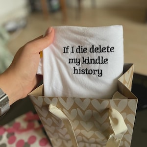 May include: White t-shirt with black embroidery that reads "If I die delete my kindle history". The t-shirt is folded and placed in a brown gift bag with white hearts.