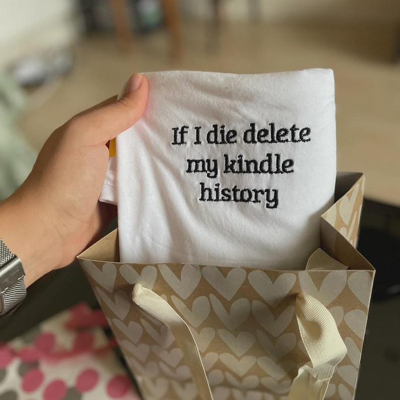 May include: White t-shirt with black embroidery that reads "If I die delete my kindle history". The t-shirt is folded and placed in a brown gift bag with white hearts.