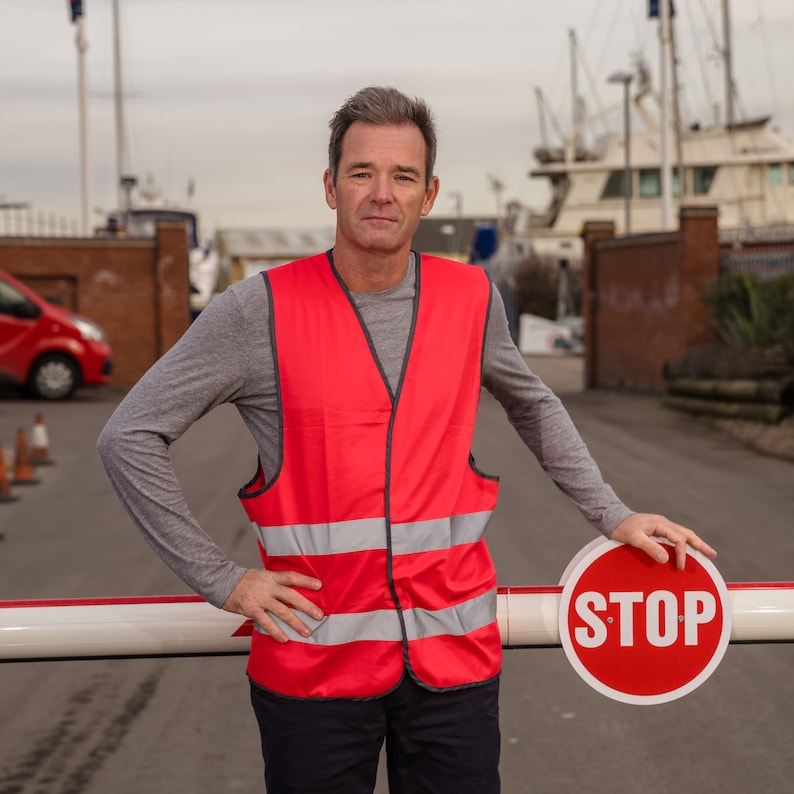 May include: A man wearing a red safety vest with reflective stripes stands in front of a white and red gate with a red STOP sign. The gate is closed.