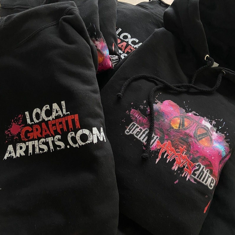 May include: Black hooded sweatshirts with white text and graphic designs. One sweatshirt has the text "LOCAL GRAFFITI ARTISTS.COM" and the other has the text "graffiti artists 2 hire" with a pink and purple graphic design.