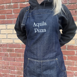 May include: A dark blue denim apron with the embroidered text "Aquila Pizza" in white. The apron features a large front pocket and adjustable straps. The apron is worn over a black hooded sweatshirt.