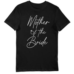 May include: Black t-shirt with white cursive text that reads "Mother of the Bride". The text includes a small ring graphic between "Mother" and "of the". A classic crew neck tee.
