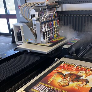 May include: A close-up of a large-format digital printer with a multi-color ink system. The printer is printing a design onto a flat surface. The design features a cartoon character with a skull and crossbones.